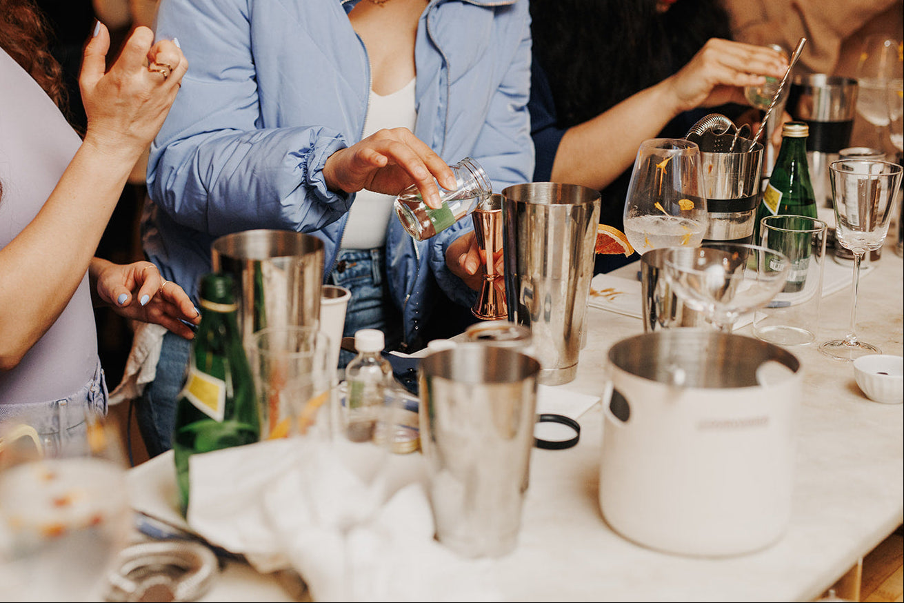 People preparing drinks at a bar with various bottles and glasses on a table.