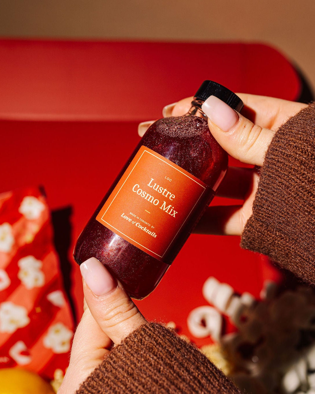 Person holding a bottle of 'Cocktail Mixer' in front of a red box with snacks.